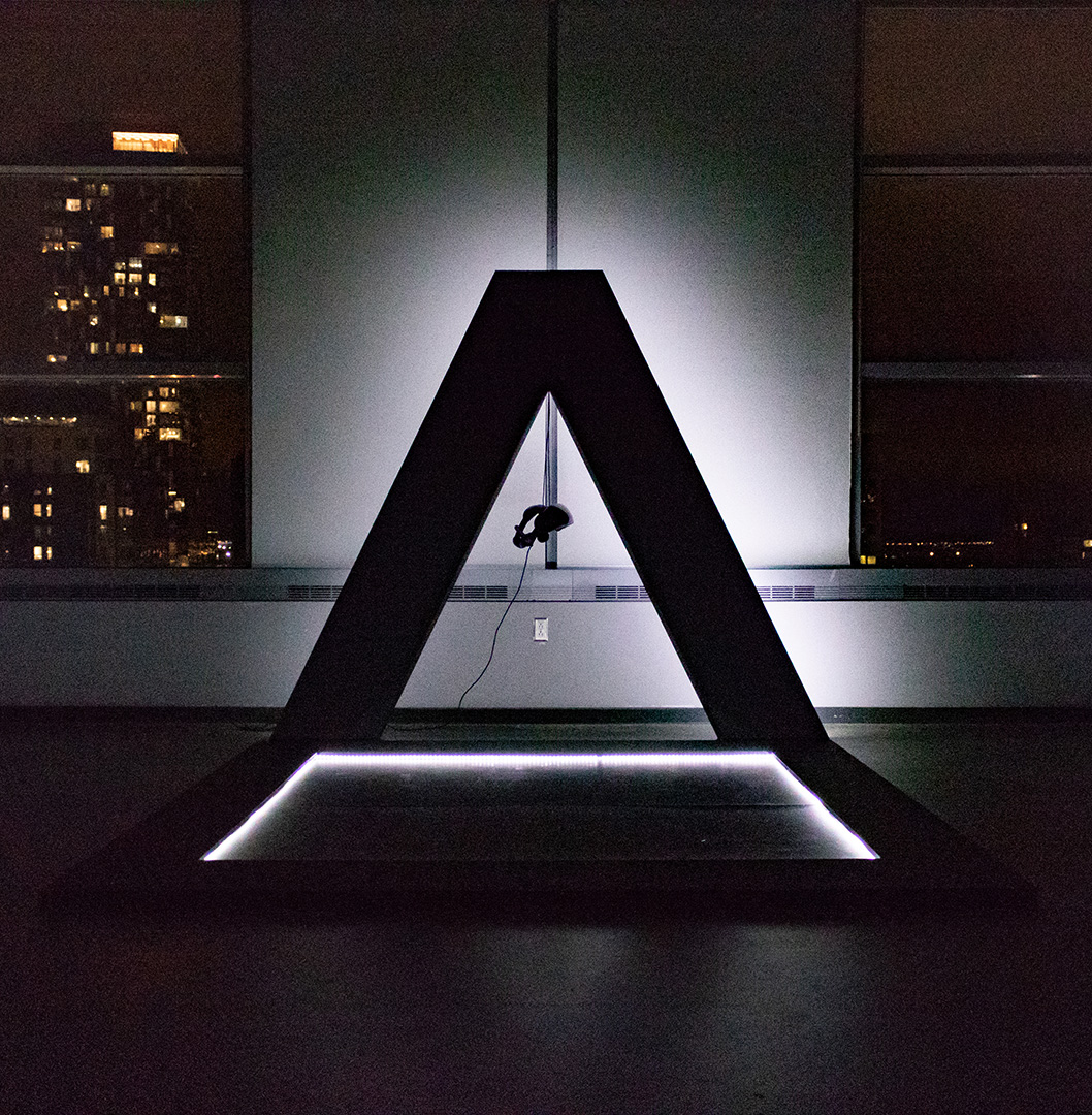 Photograph of an installation sculpture shaped like a triangle with a light behind it in a dark room.
